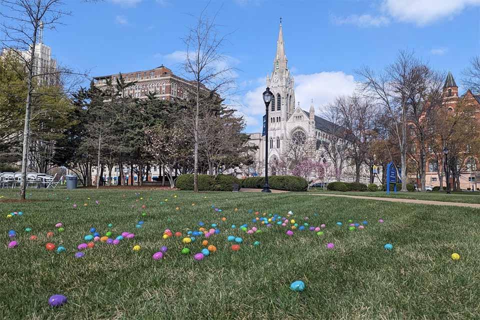 在一个阳光明媚的日子里，五彩缤纷的复活节彩蛋散落在绿色的草坪上，背景是大学教堂和杜堡大厅