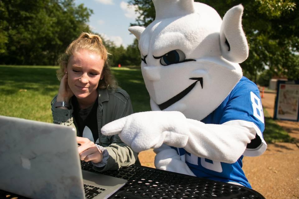 The Billiken working on a laptop in the quad with a student.