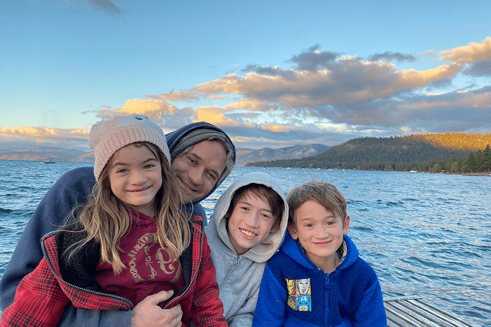 日落时分，特拉维斯·霍夫斯泰特和他的三个孩子穿着夹克，戴着帽子，在远处群山环绕的一片水域前摆姿势.
