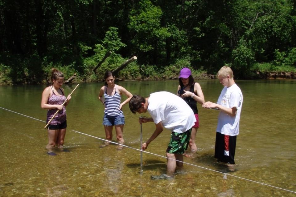 学生们站在与膝盖齐平的河里，拿着设备看着河水.