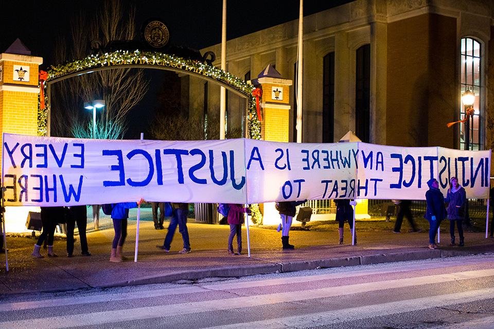 Students stand along a street on campus in front of an archway decorated by Christmas lights. They hold a long banner that reads "Injustice Anywhere is a Threat to Justice Everywhere"