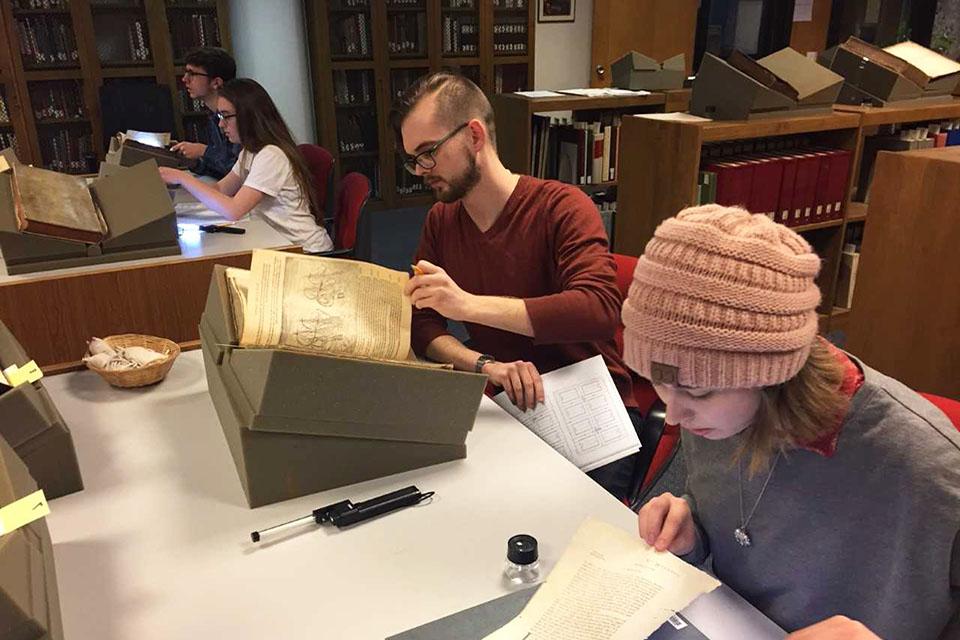 Students sit at tables in a library, examining old texts.