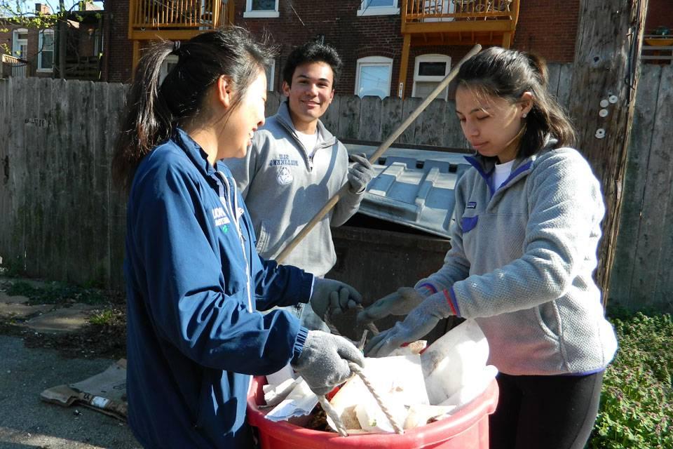 Two students wearing work gloves examine the contents of a bucket while another student holds a work tool in the background.