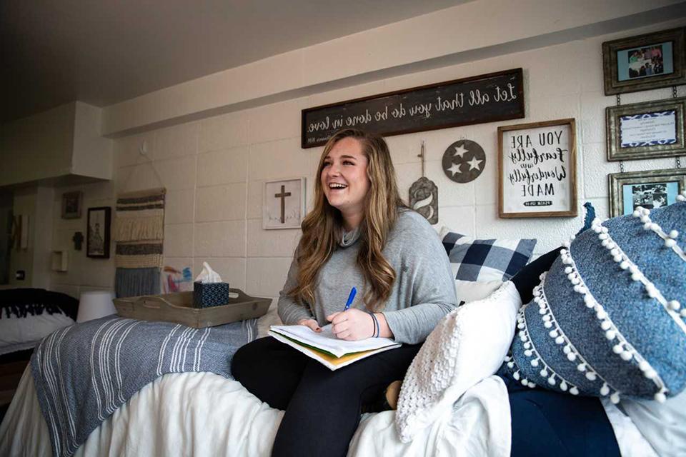 A student sits on her bed in a dorm room, laughing while writing in a notebook.