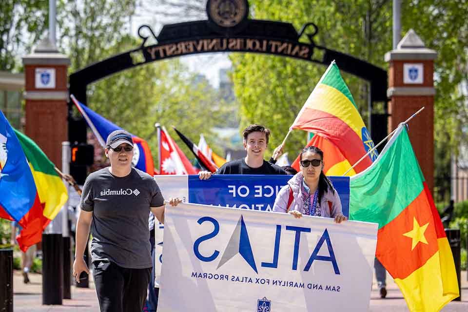 Students holding a sign reading Sam and Marilyn Fox Atlas Program in front of a parade of flags