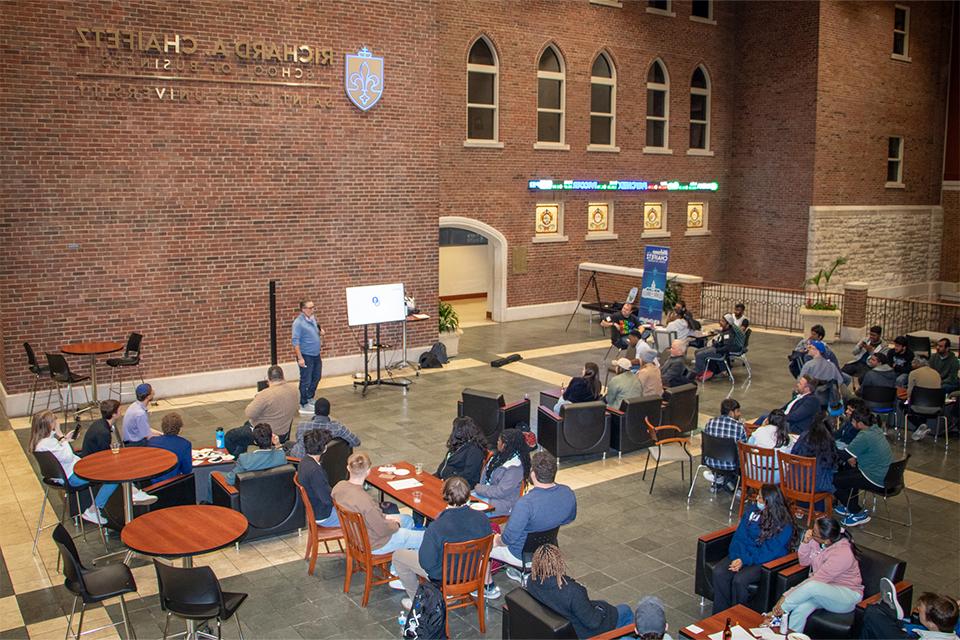 People sit around tables in an atrium while a speaker holds a microphone at one end of the room.