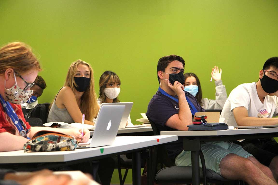 Students in masks sitting in a classroom listening to a lecture; one student raises her hand.