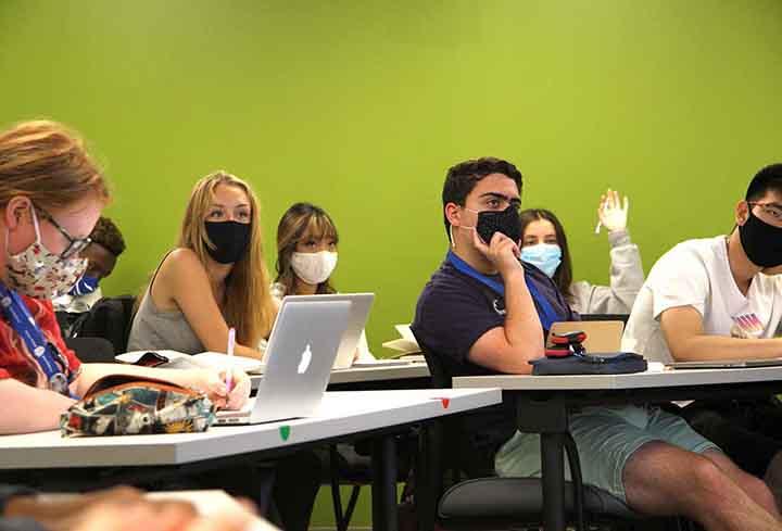 学生们坐在教室的桌子上，戴着口罩向前看. 一个学生举起了手.