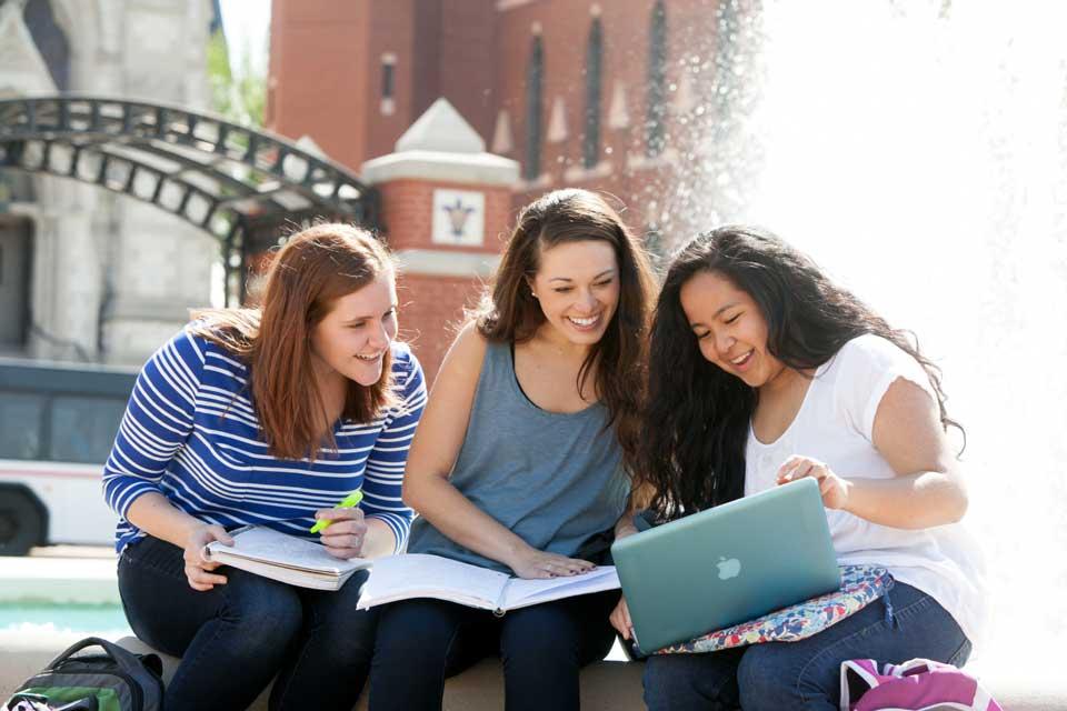 三个学生共用一台笔记本电脑, 膝上还放着笔记本, 坐在喷泉前的时候.