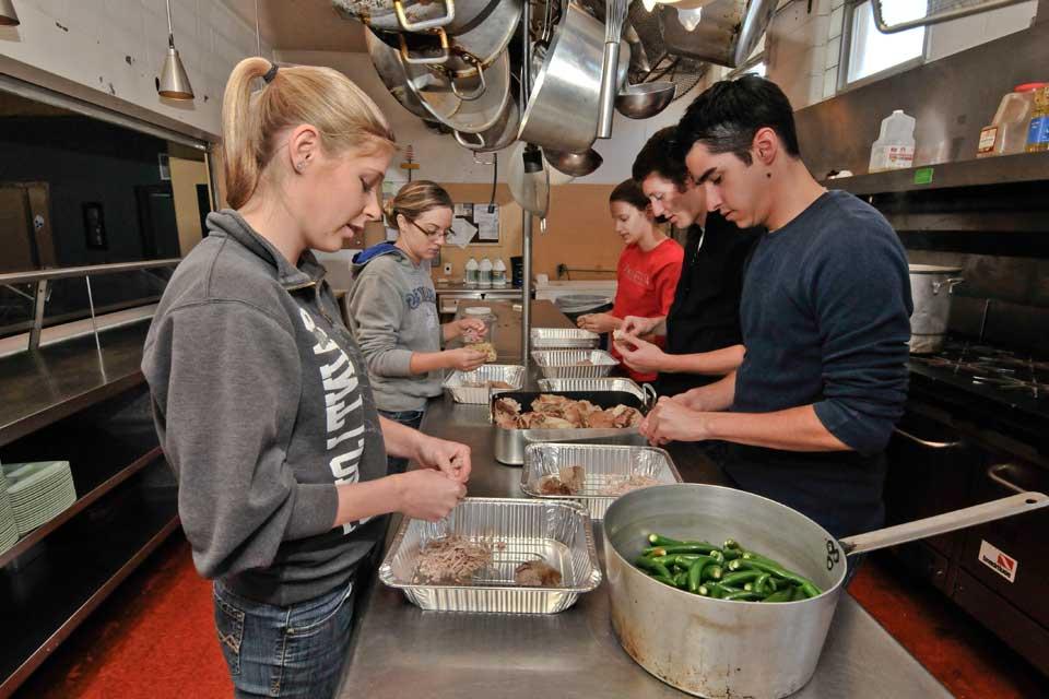 Students standing at a counter in the middle of a  a commercial kitchen, preparing food.