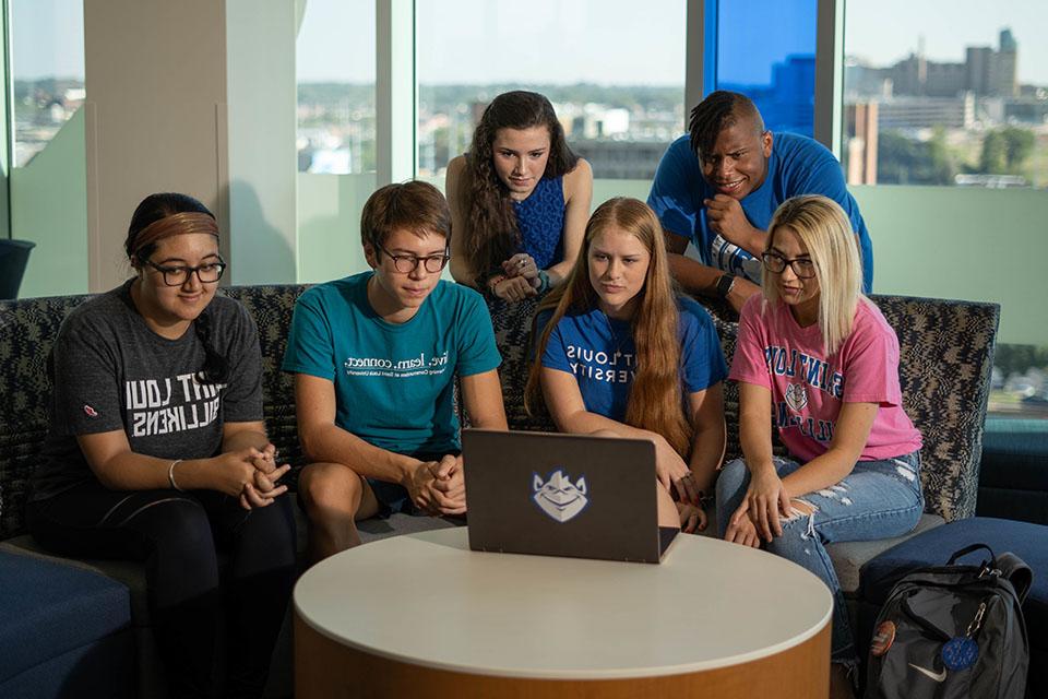 Four students sit on a semi-circular sofa in a residence hall lounge, two stand behind them. All looking at a laptop screen.