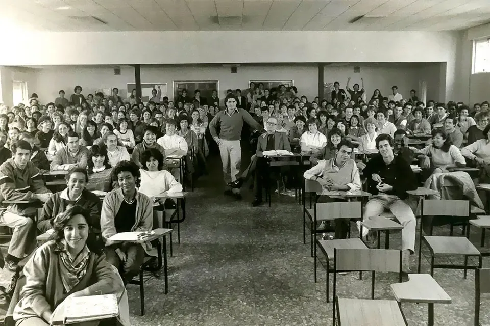 A black and white photo features students sitting at classroom desks while a professor stands in the center of the room.