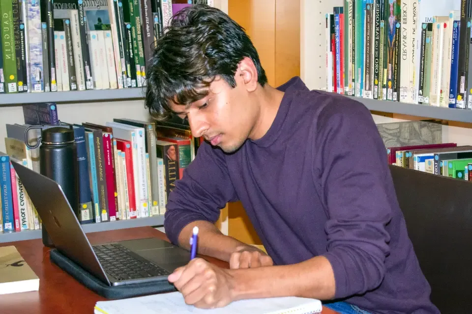 一个学生坐在桌子旁，用笔记本电脑在笔记本上写东西.
