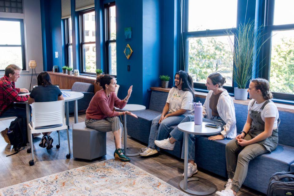 Students and a faculty member sit on chairs and benches in a room with windows and plants.