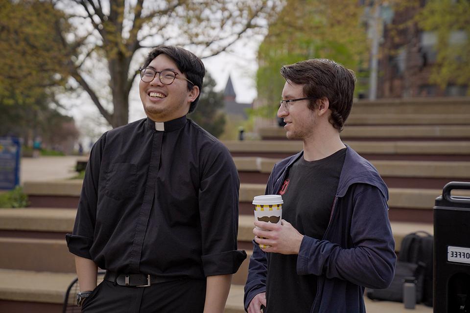 A priest stands next to a man attending Java with the Jesuits. 他们边说边笑.