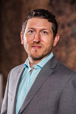 MOCRA Director David Brinker, 穿着浅蓝色的衬衫和灰色的运动外套, 在一个抽象的棕色背景前摆姿势