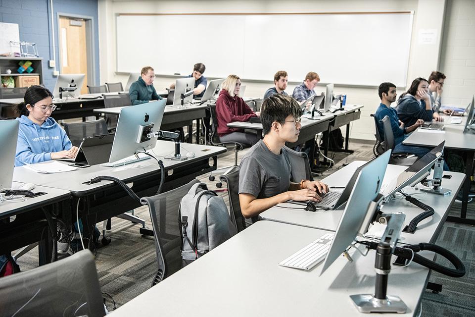 学生们坐在教室里的课桌前，拿着笔记本电脑和电脑显示器.