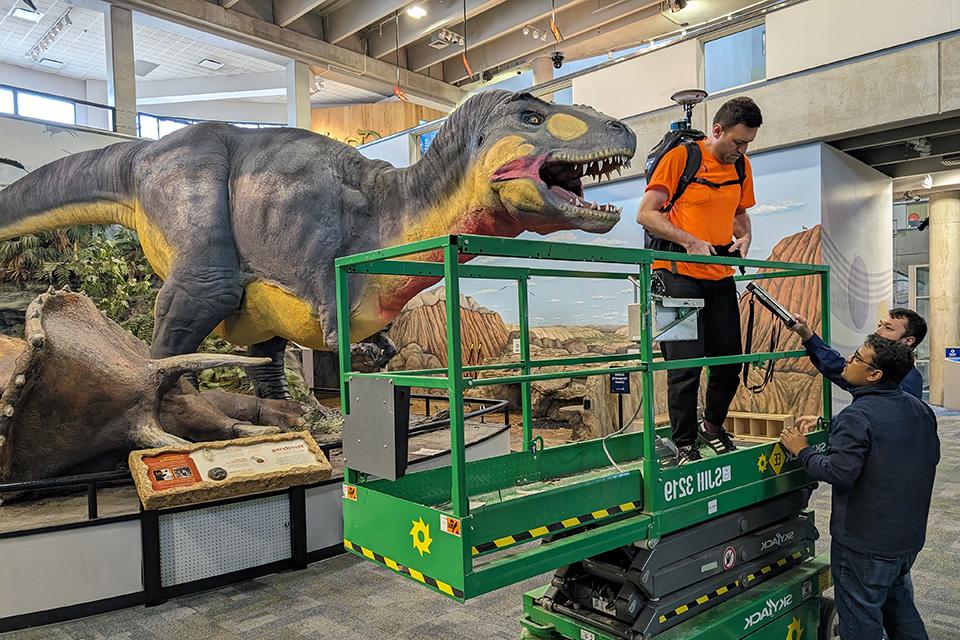 一个学生站在电梯里，背上背着一件大设备，站在一个乳胶恐龙前面.