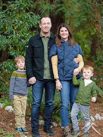 Tim Bantle's family portrait. Two young boys in sweaters flank Sara, who wears a navy quilted jacket and jeans, and Tim, who wears a green shirt, black jacket, and dark jeans. They stand in a forest setting.